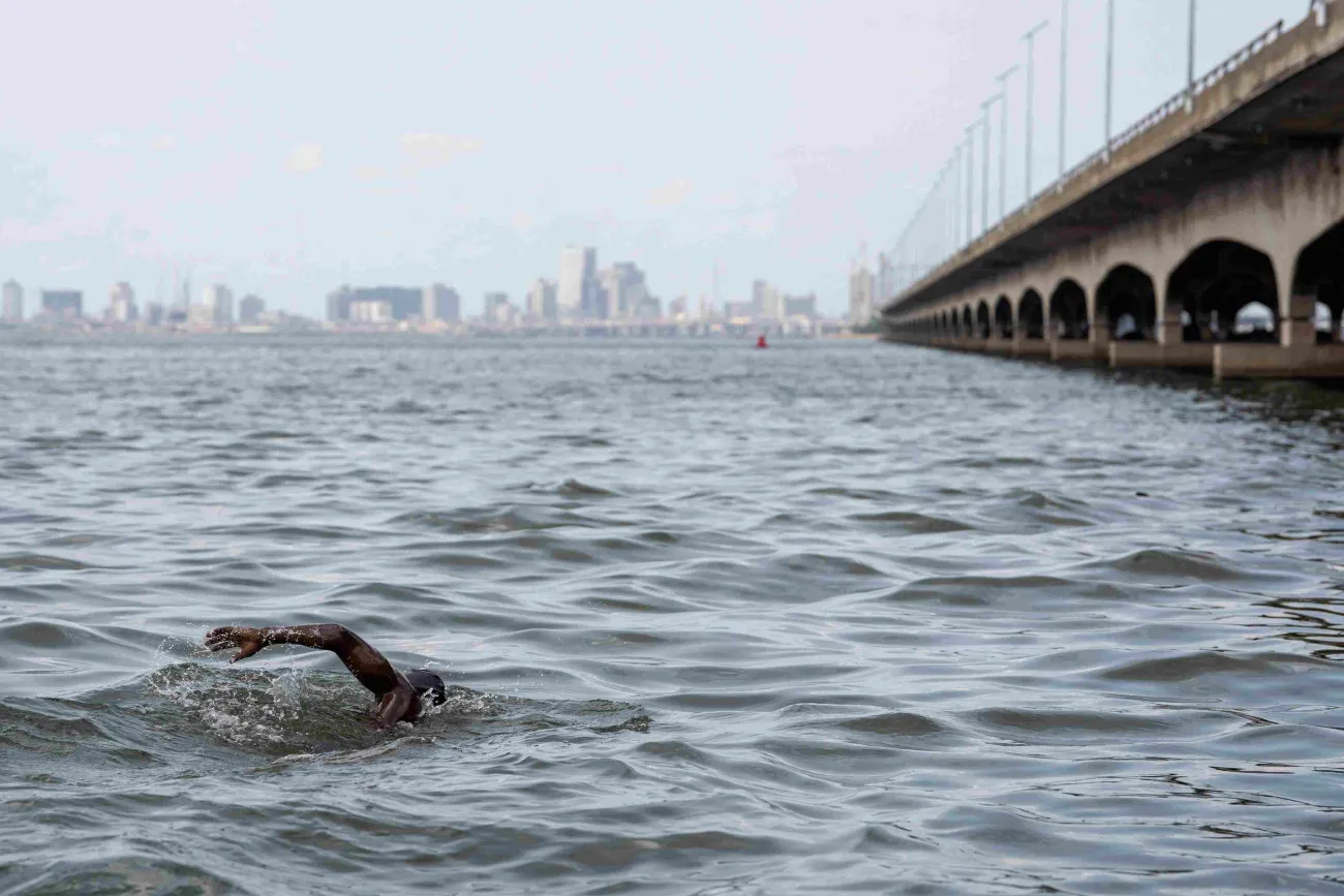 A person swimming in a large body of water alongside a bridge, heading towards a city on the horizon.