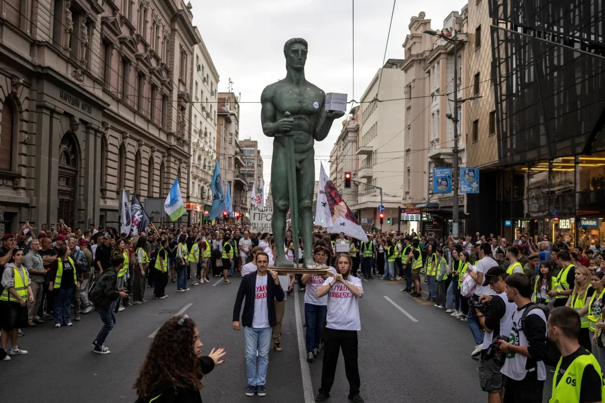 A large street demonstration in a city centre, with two protesters carrying a towering bronze statue through a dense crowd. Many participants wear fluorescent yellow vests and carry flags and banners, while spectators line both sides of the street.