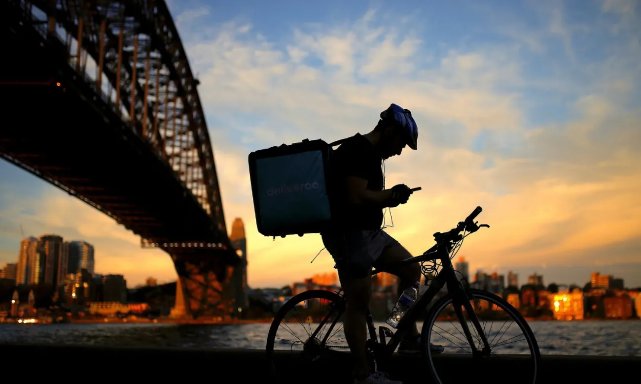A delivery man checks his phone as he rests on his bicycle under the Sydney Harbour Bridge at sunset in Sydney